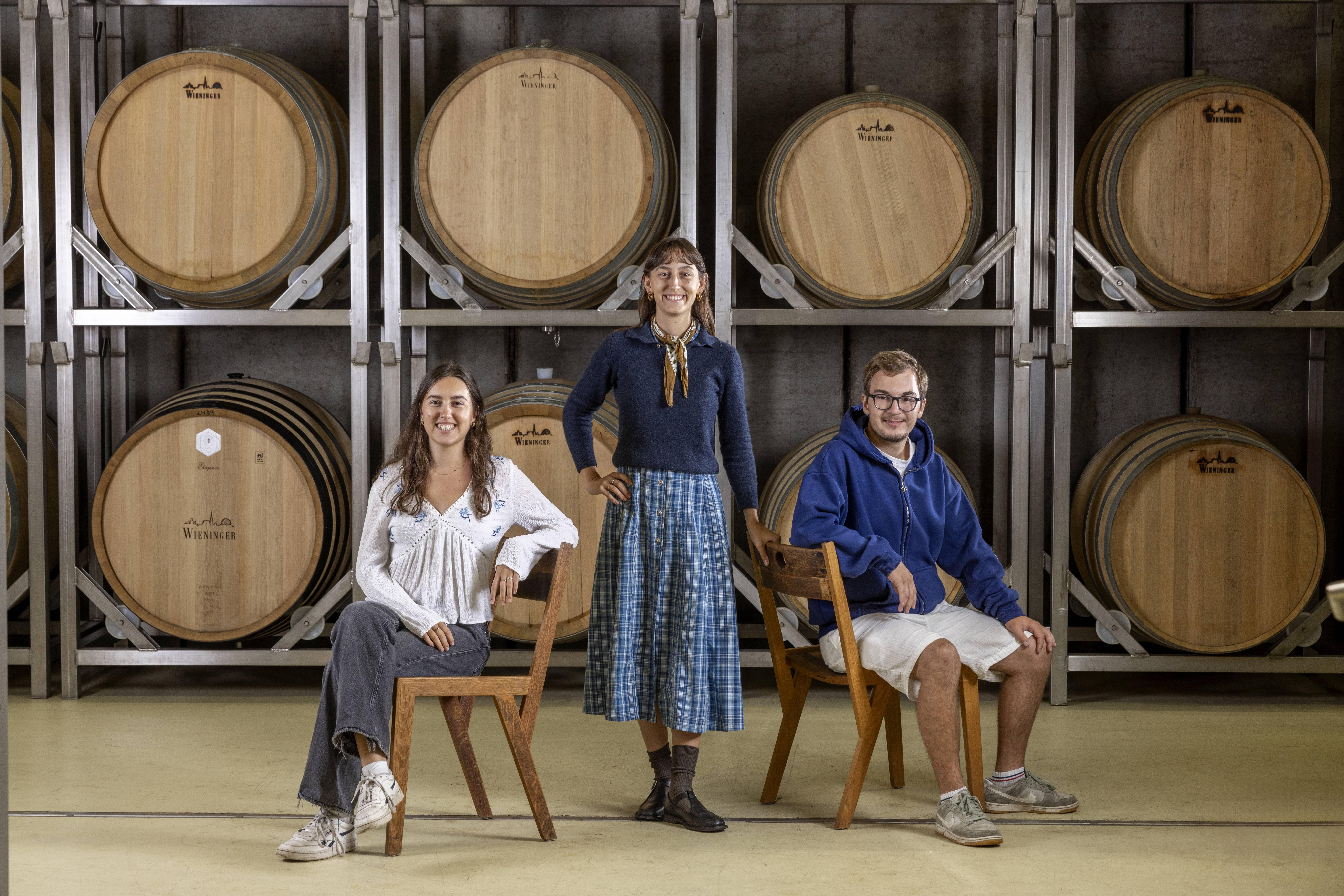 Das Foto zeigt Katharina (links sitzend), Sophie (mittig stehend) und Maximilian Wieninger (rechts sitzend) wie sie fröhlich im Barrique-Weinkeller vor den großen Holzfässern posieren.